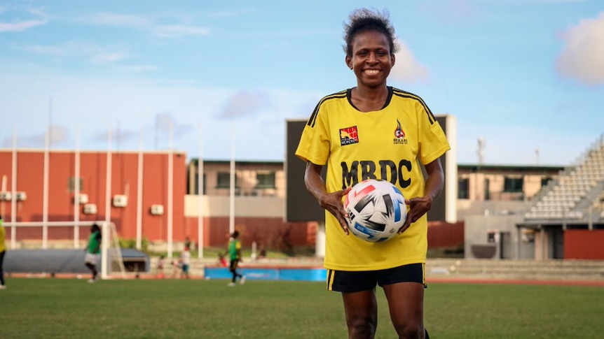 A female soccer player smiles broadly as she holds a ball in her hands and poses for a picture standing on a football pitch.