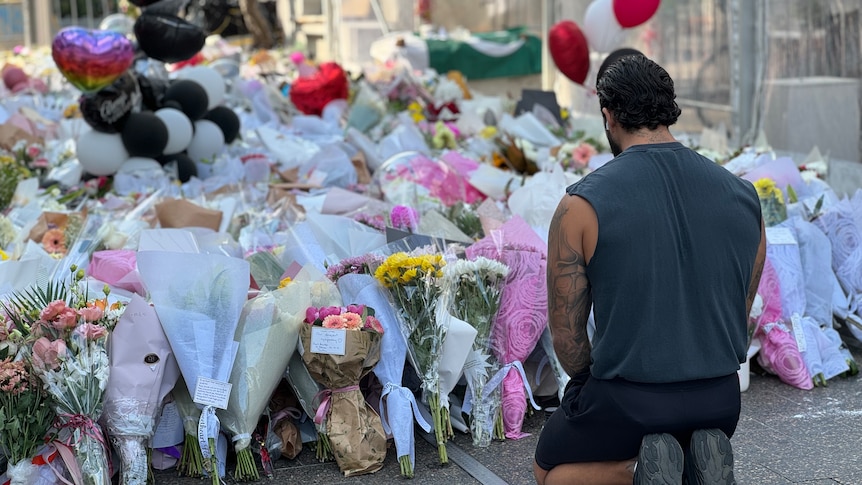 A man sitting looking at flowers in a tribute at Bondi Junction shopping centre.