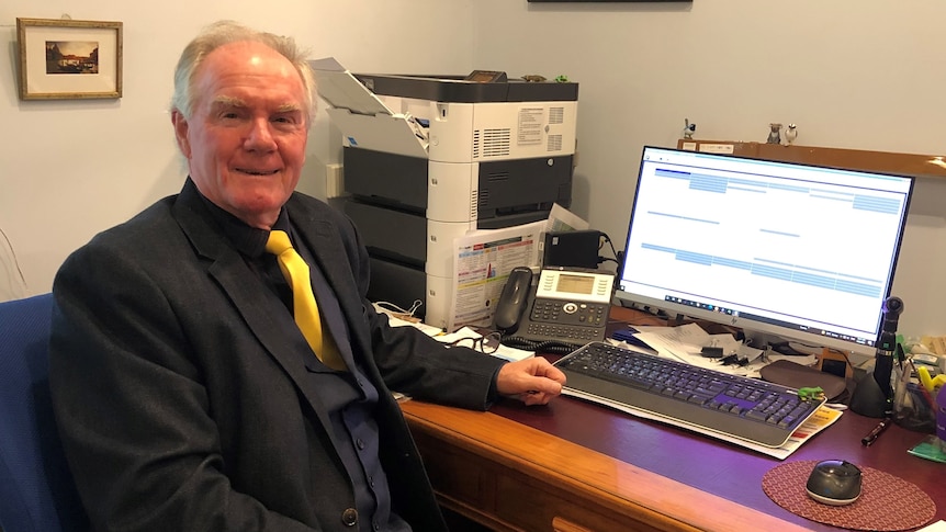 An older man in a black suit, vest and yellow tie sits at a desk with an open laptop on it