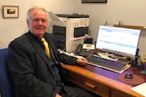 An older man in a black suit, vest and yellow tie sits at a desk with an open laptop on it