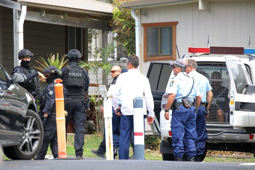 Police officers stand around near a police car with a man inside.