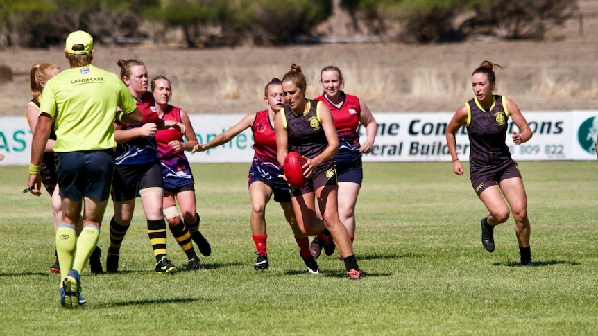 A woman footballer in a brown and yellow jumper holds the ball ahead of a pack of women players.