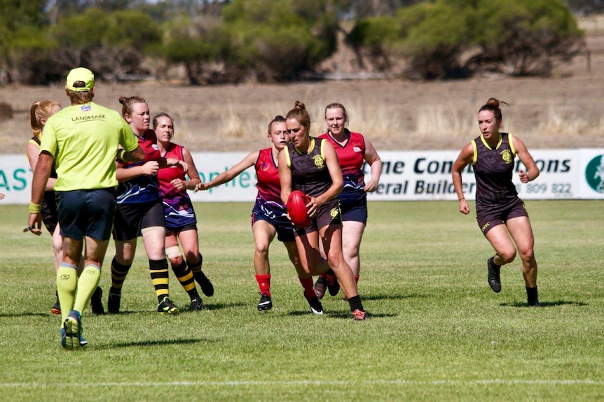 A woman footballer in a brown and yellow jumper holds the ball ahead of a pack of women players