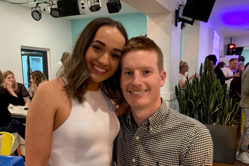 Hannah wearing a white dress and her partner wearing a button up shirt, in a hall with other people, smiling at the camera.