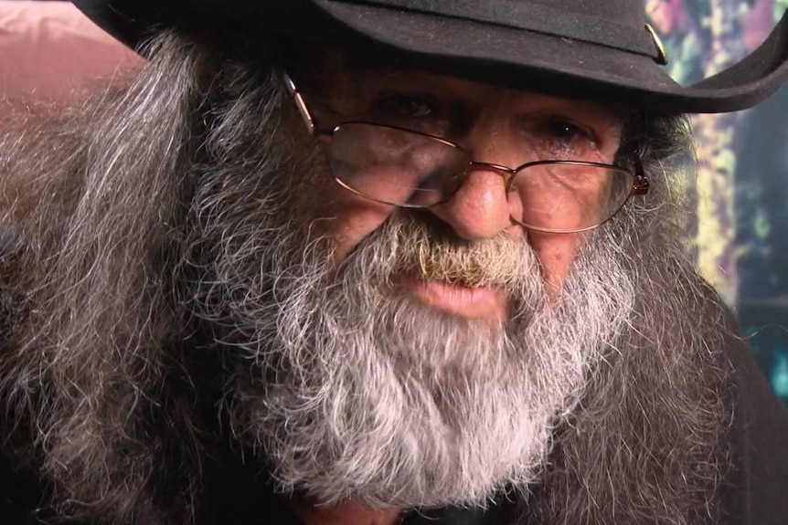 A tight head shot of an Indigenous man with a beard wearing a black hat and spectacles.