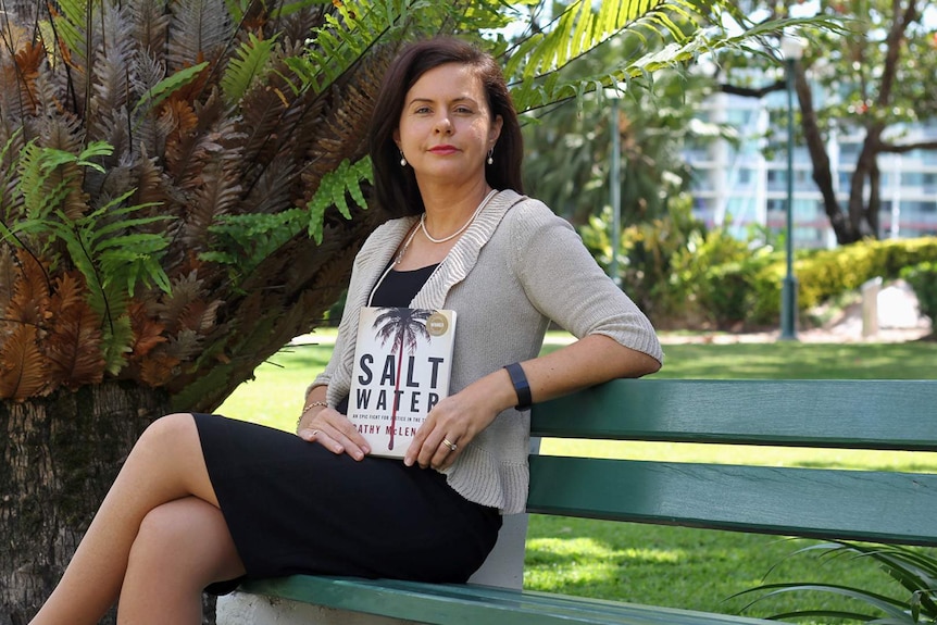Author Cathy McLennan is seated on a park bench holding a copy of her book Saltwater