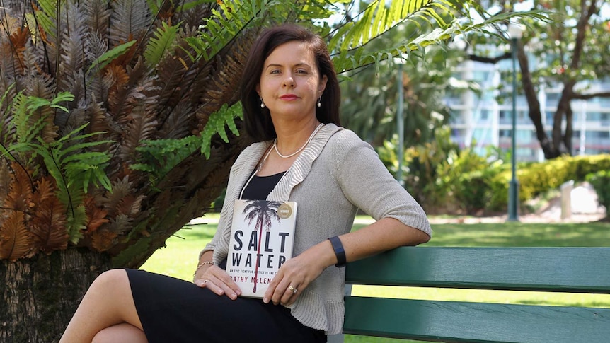 Author Cathy McLennan is seated on a park bench holding a copy of her book Saltwater