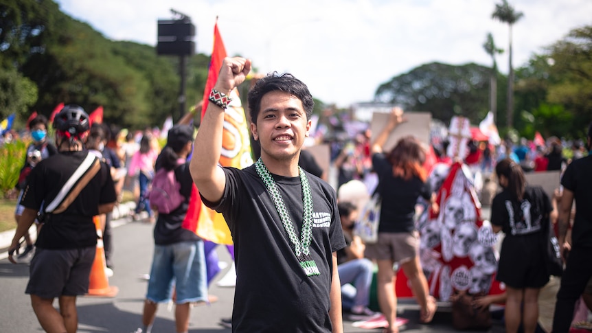 Young man stands in front of a rally holding up a fist. 