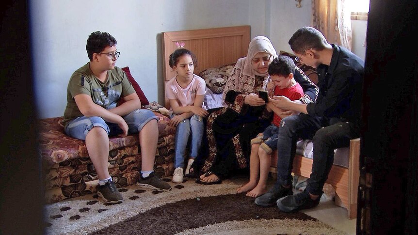 Amal Al Halabi and her grandchildren sit in a bedroom looking at photos on a mobile phone.