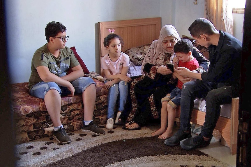 Amal Al Halabi and her grandchildren sit in a bedroom looking at photos on a mobile phone.