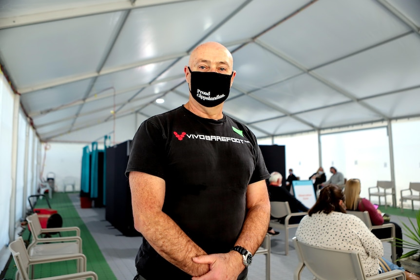 Man wearing black t-shirt and black mask stands arms together inside marquee with people and booths behind him