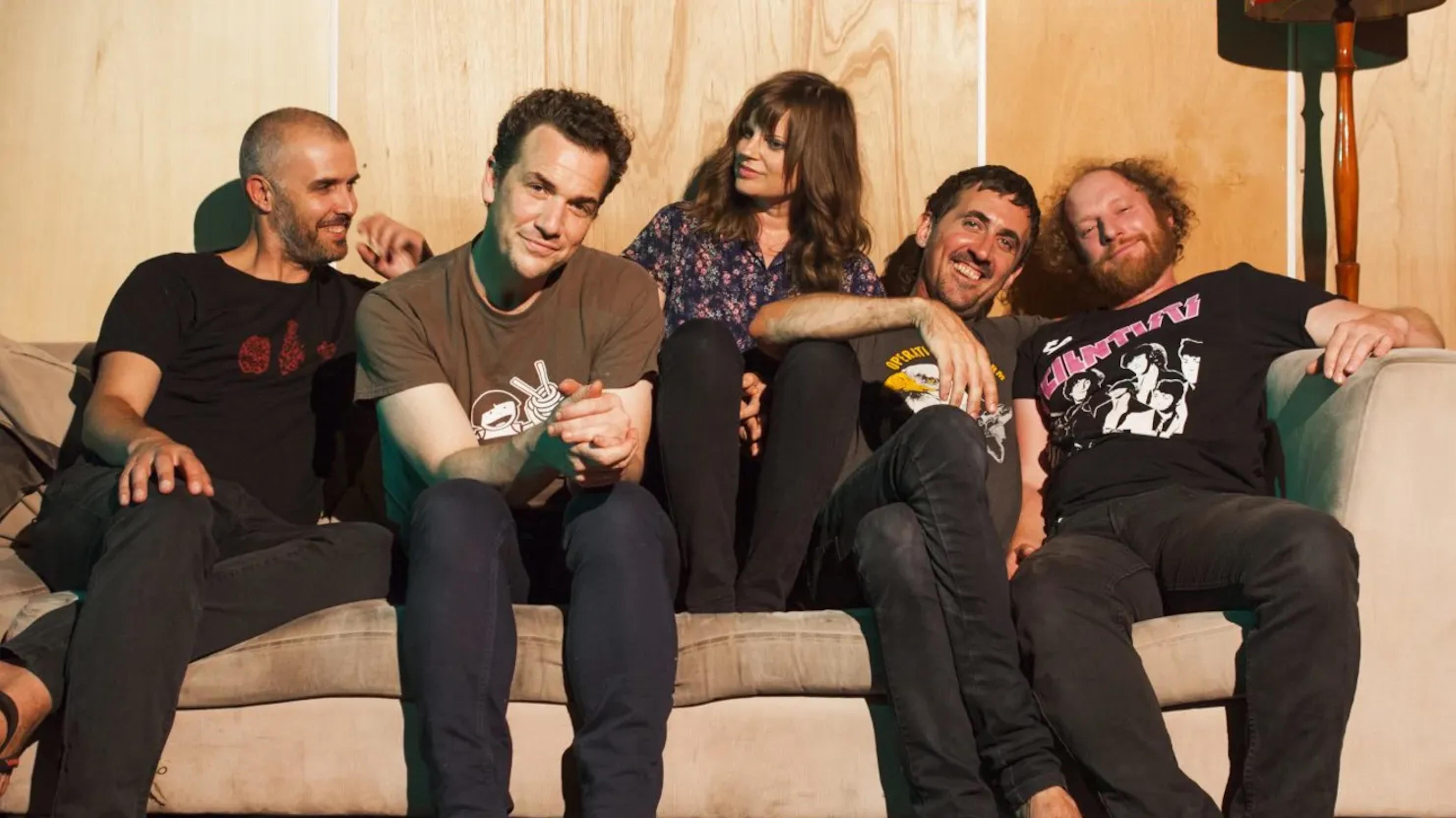 Five people sitting close together on a couch in casual clothing, against a wooden wall indoors.