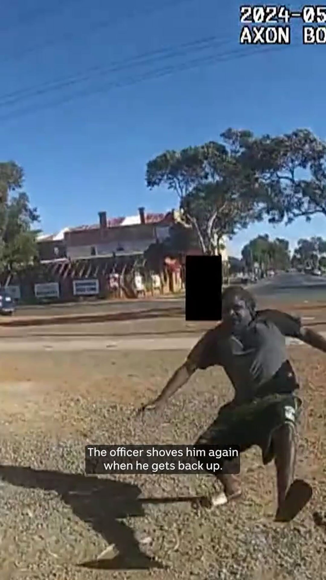 A man with dark-tone skin stumbles with one foot up on a rural street. A black rectangle obscures part of the frame