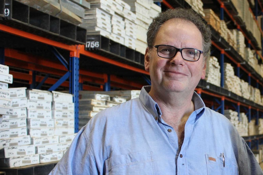 A middle-aged man in a blue shirt and glasses stands in front of a warehouse shelf