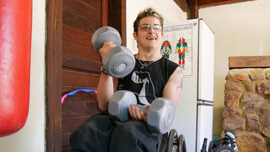 A man in a wheelchair holding two dumbbells in his gym with a boxing bag in the foreground.