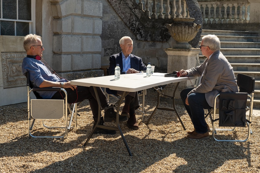 Three elderly white men sit in suits chatting around a trestle table outside a sandstone house.