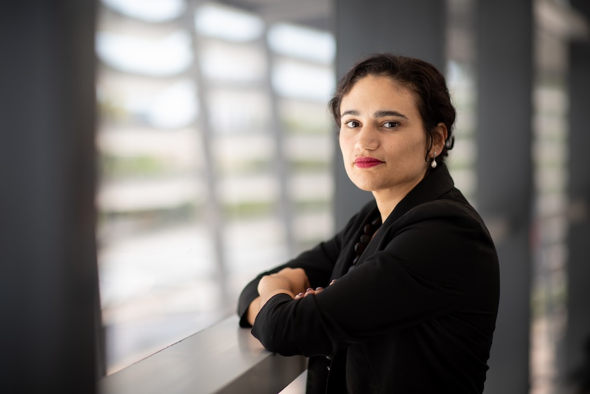 A serious-looking woman looking at the camera while crossing her arms and leaning on a balcony railing. 