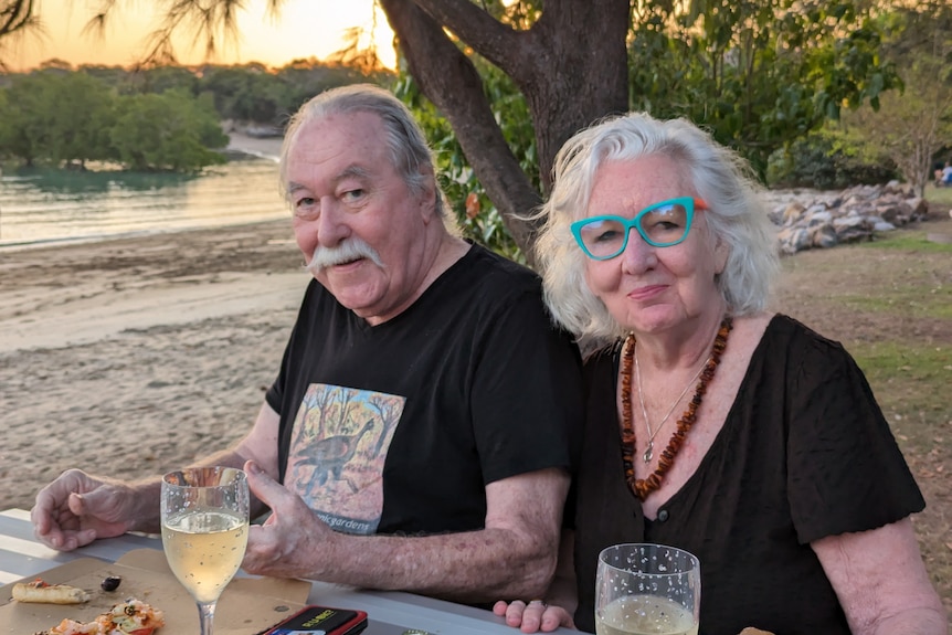 An older couple sit at an outdoor table by the beach with glasses of wine and pizza