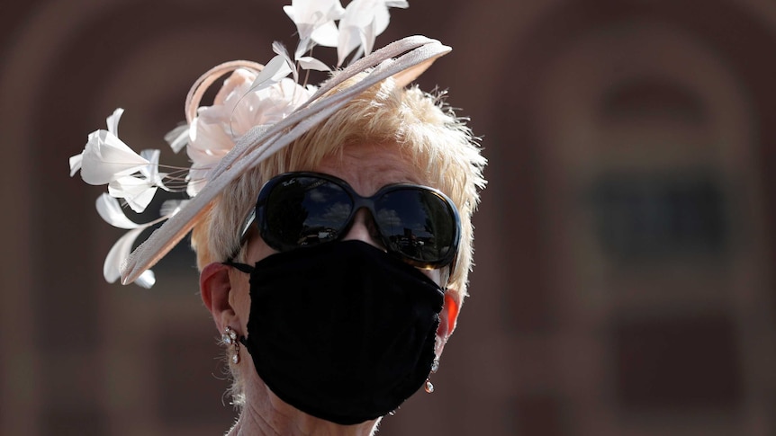 A woman with blonde hair wearing a fascinator, sunglasses and a black mask.