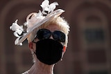 A woman with blonde hair wearing a fascinator, sunglasses and a black mask.
