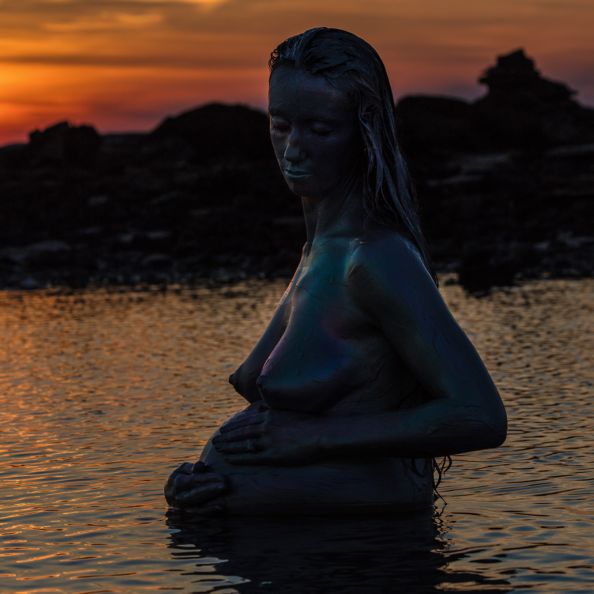 Colour square cropped image of a nude pregnant woman clutching her stomach and standing in water as the sun sets.