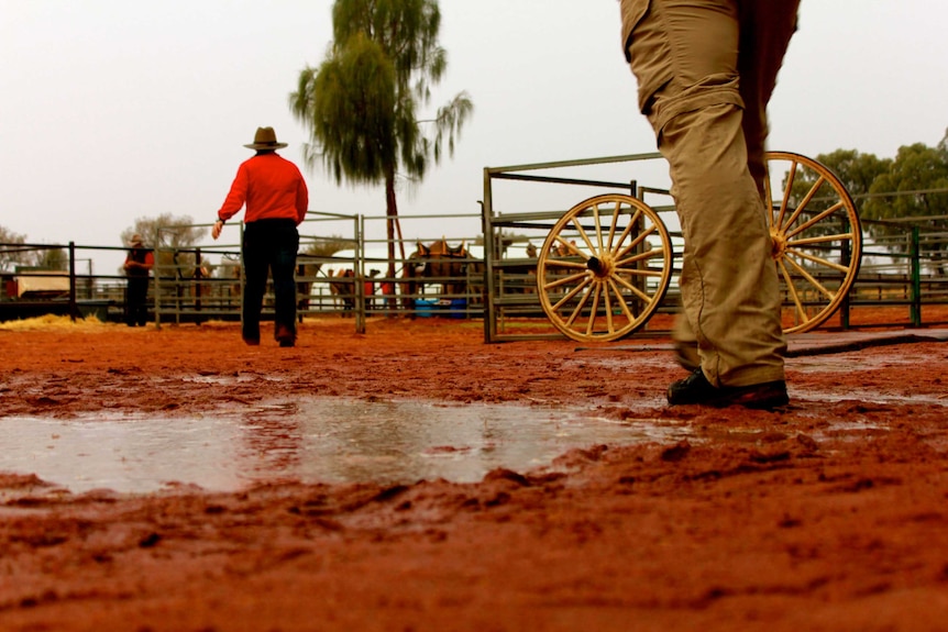 A picture of a red dirt puddle at foot level. One person's legs as they walk is the foreground and another in the background.