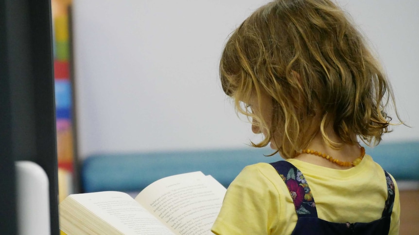 A close-up of a young girl reading a book. The young girl has dark brown, shoulder length hair.