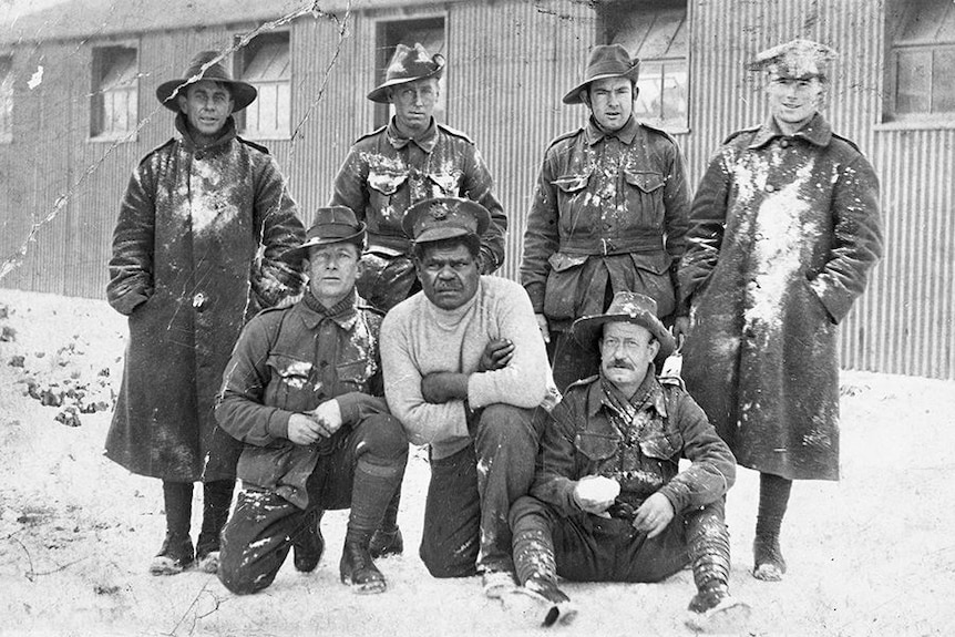An Aboriginal man wearing a grey shirt surrounded by other men in army uniform standing in snow