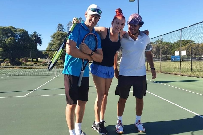 A smiling tennis player stands on court, putting her arms around two coaches either side of her.