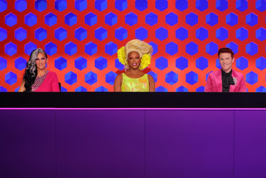 A pane of a TV, with RuPaul in drag, Rhys Nicholson in a pink jacket and Michelle Visage