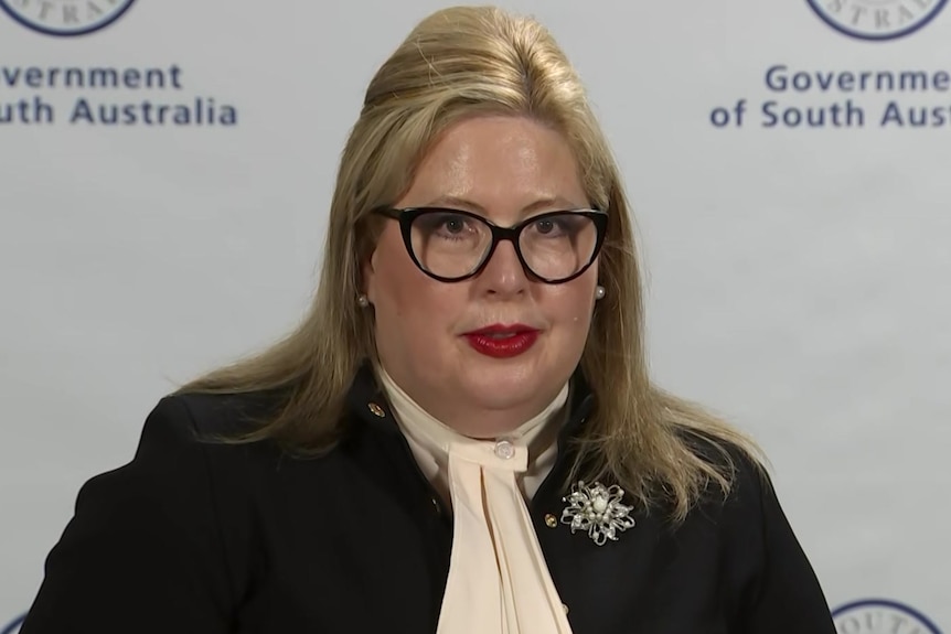 A woman with blonde hair wearing glasses, a blush top, a black jacket and a brooch in front of a banner