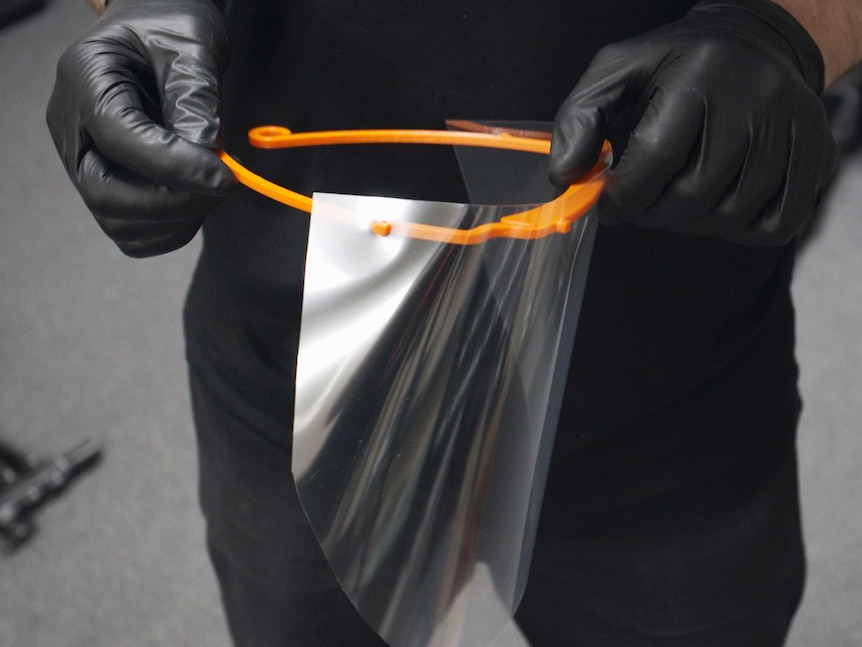 A man wearing gloves holds a plastic face shield