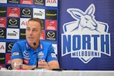 A man with close-cropped hair sits behind a microphone in front of a North Melbourne banner and advertising logos.