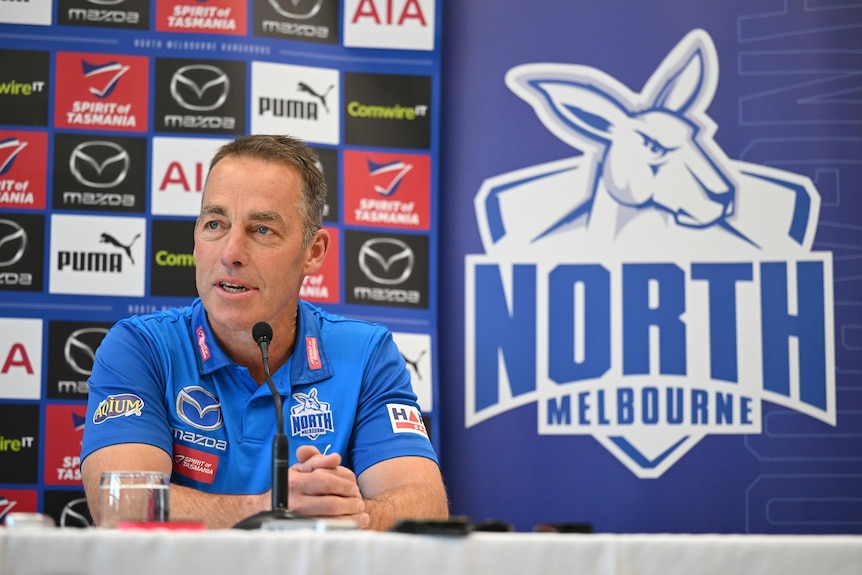 A man with close-cropped hair sits behind a microphone in front of a North Melbourne banner and advertising logos.
