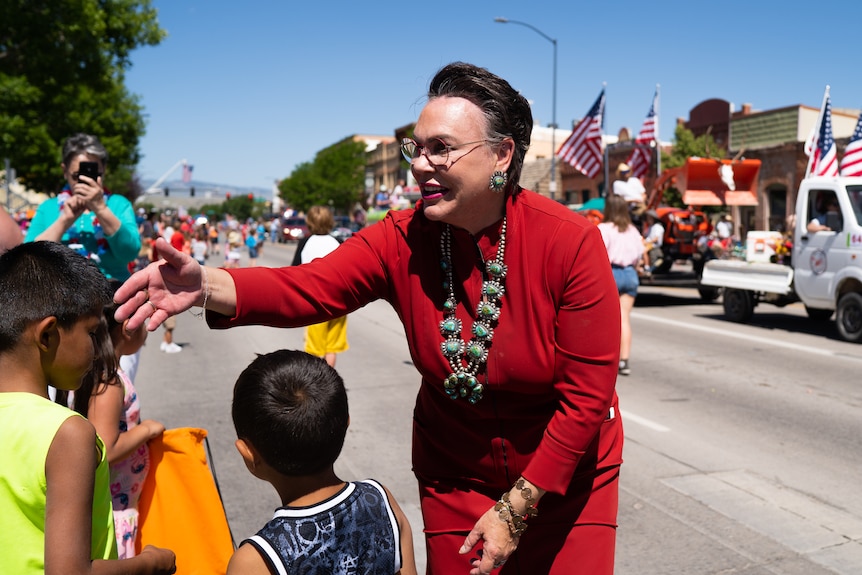 A woman with close cropped hair and a red dress shakes hands with children on a street 