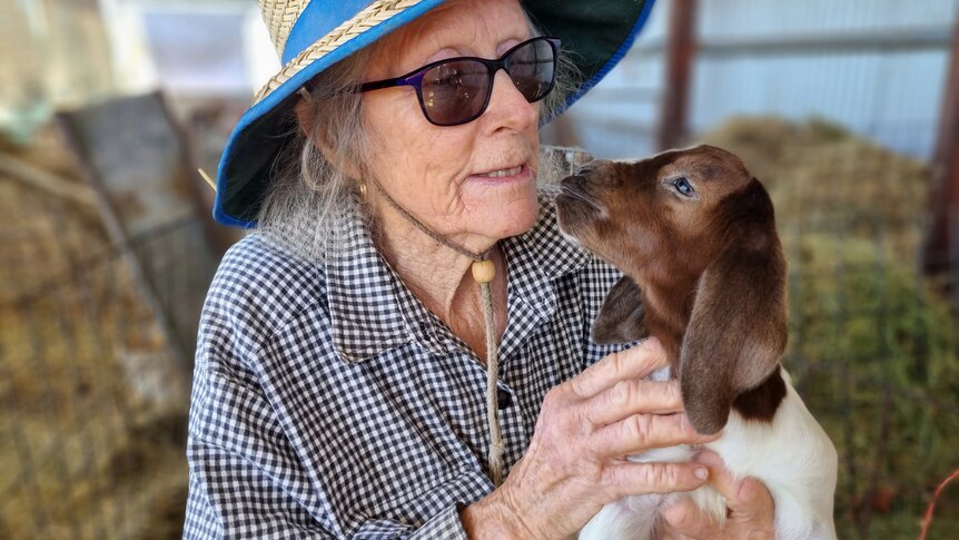 A woman in a hat and glasses holds a goat.