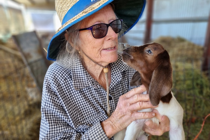 An elderly woman in a hat and glasses hold a goat.