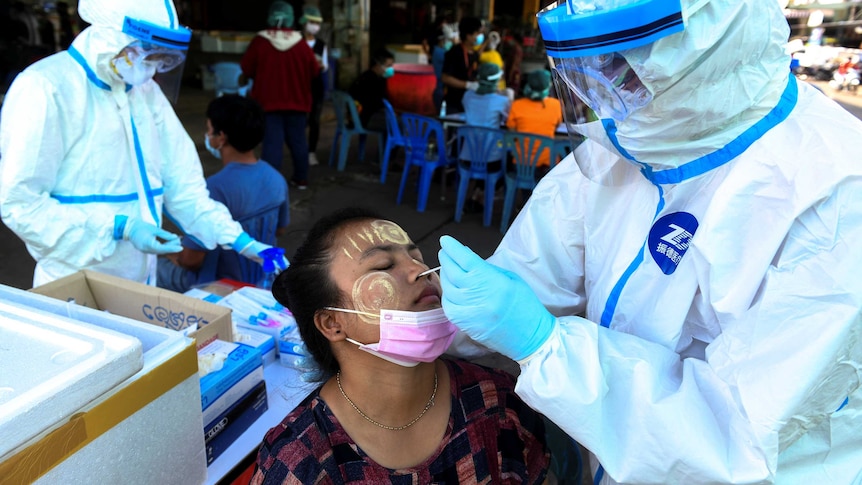 A woman with traditional yellow smeared on her face leans her head back for a nasal swab from a person in a protective suit.