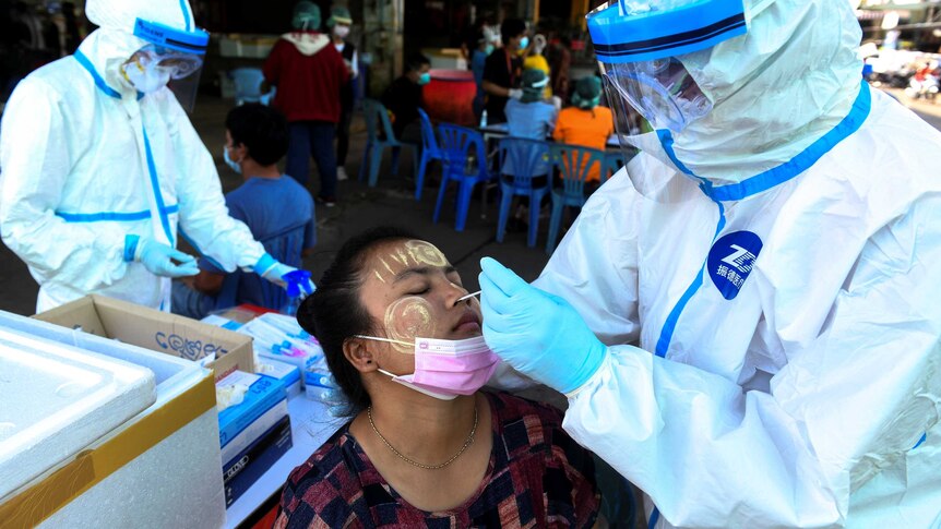 A woman with traditional yellow smeared on her face leans her head back for a nasal swab from a person in a protective suit.