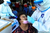 A woman with traditional yellow smeared on her face leans her head back for a nasal swab from a person in a protective suit.