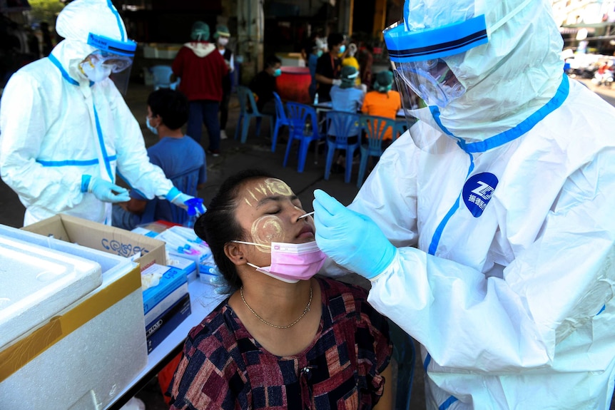 A woman with traditional yellow smeared on her face leans her head back for a nasal swab from a person in a protective suit.