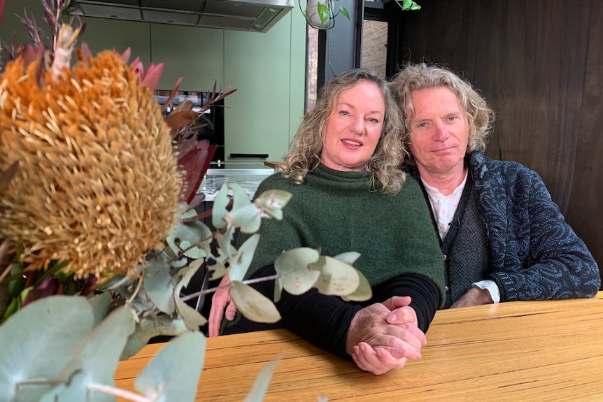 A woman and man with blonde hair sit next to a floral arrangement