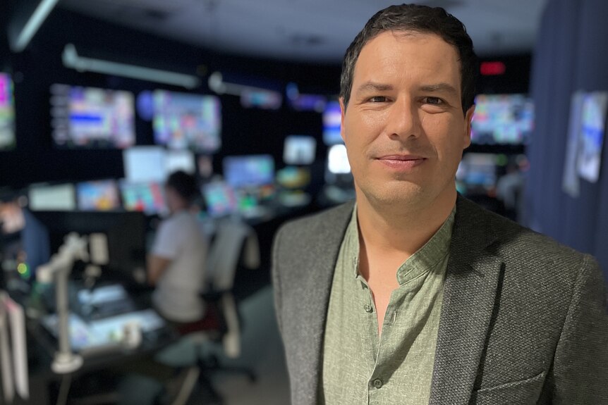 French journalist Guillaume Pitron in a grey jacket and green shirt. He is in a TV control room with many screens behind him.