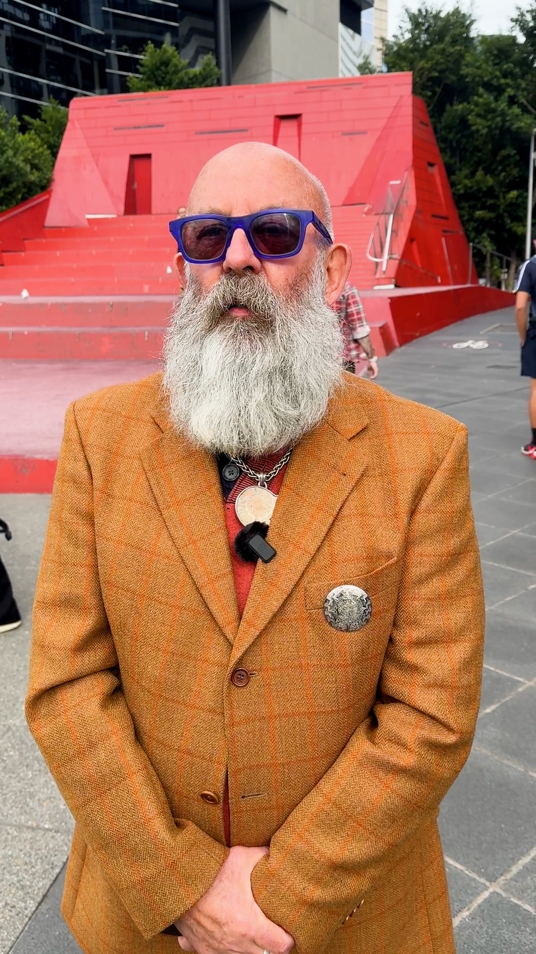 Un hombre barbudo con gafas de sol azules se encuentra frente a un alto edificio rojo.