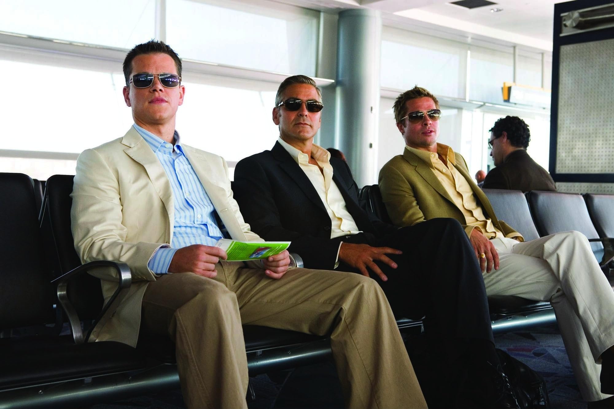 Brad Pitt, George Clooney, and Matt Damon sitting on chairs in an airport in Oceans 13. 