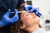 A woman lies back in a chair smiling, while someone injects her forehead with a syringe.