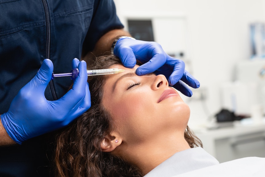 A woman lies back in a chair smiling, while someone injects her forehead with a syringe.