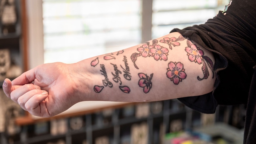 A woman's arm with pink flowers and the words 'goddess, angel, warrior' tattooed on it.