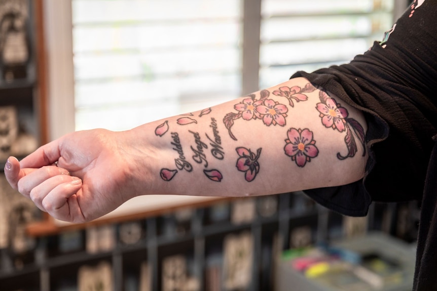 A woman's arm with pink flowers and the words 'goddess, angel, warrior' tattooed on it.