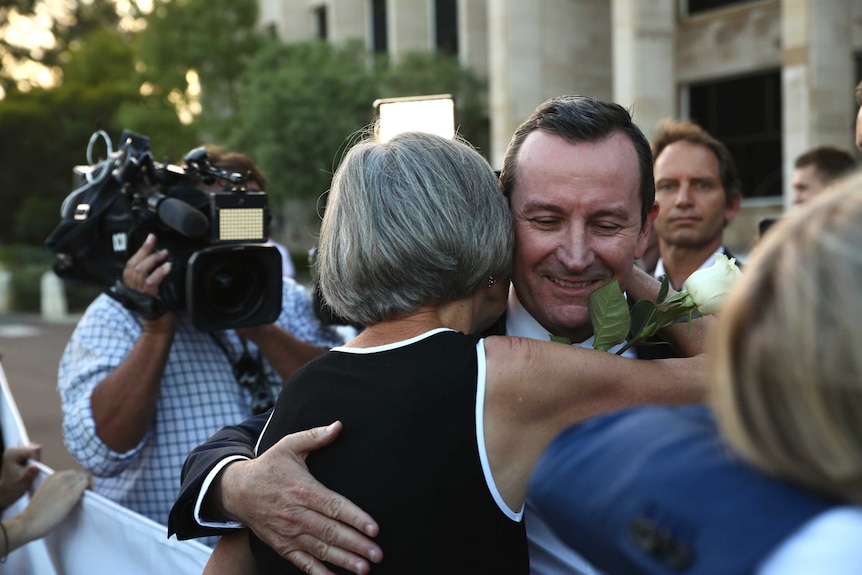 A woman smiles and embraces a man while holding a sign on the steps of parliament